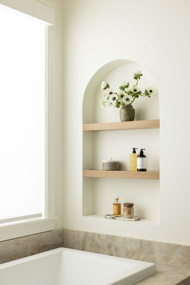 a bath tub sitting under a window next to a shelf filled with bottles and soaps