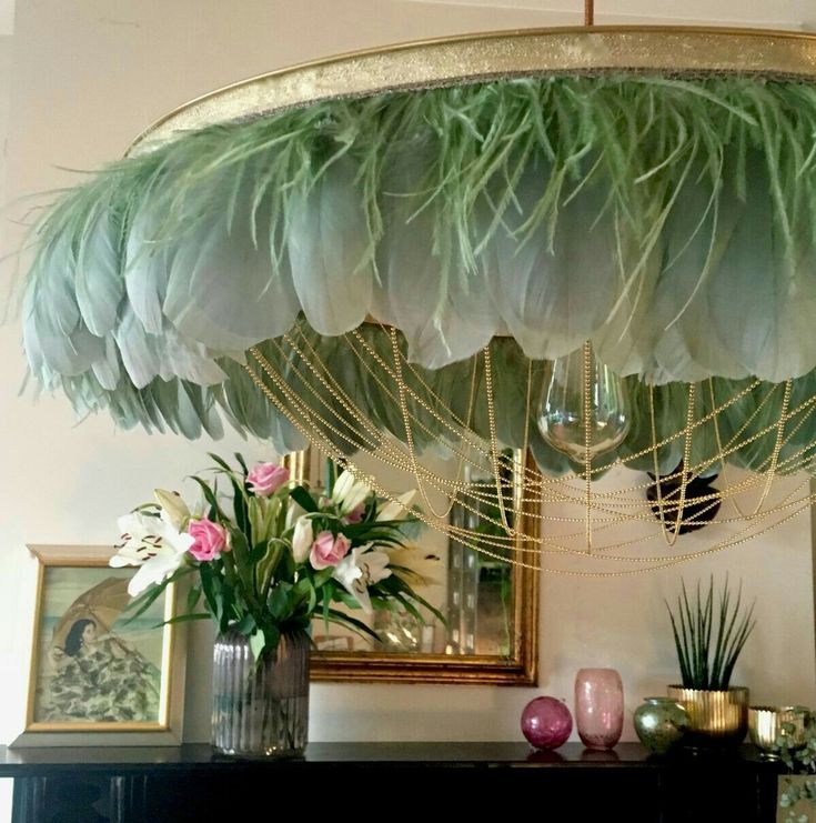 a lamp shade with green feathers hanging from it's ceiling above a table filled with vases and flowers