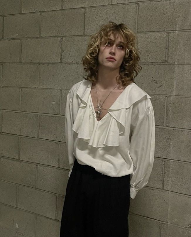 a woman standing in front of a brick wall wearing black pants and a white blouse