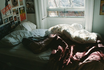 an unmade bed in front of a window with pictures on the wall behind it