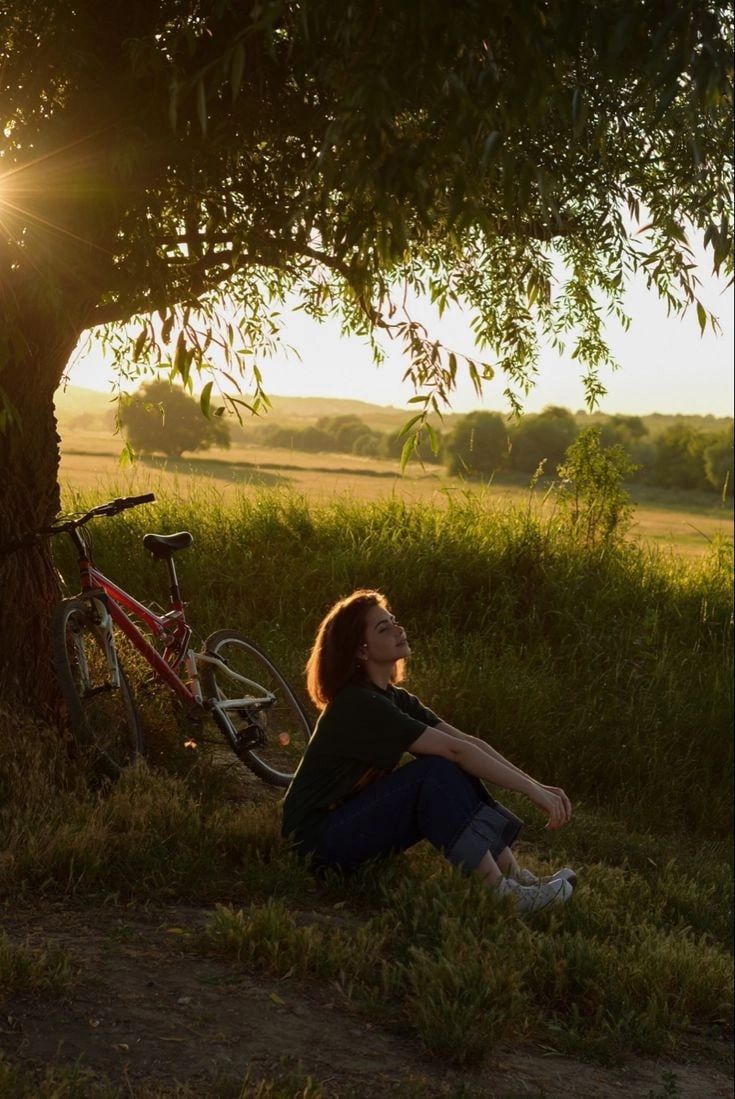 a woman sitting in the grass next to a bike under a tree with the sun shining behind her