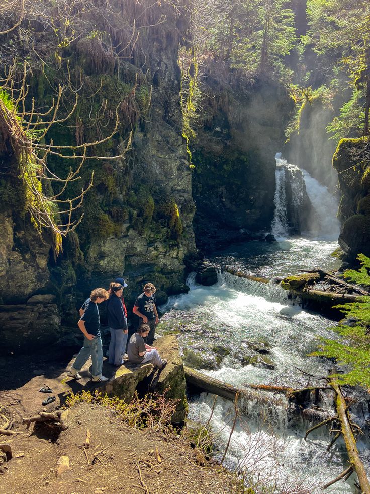 three people standing on the edge of a cliff next to a river with a waterfall in the background