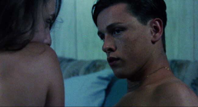 a man with no shirt standing next to a woman in bed looking at him from behind