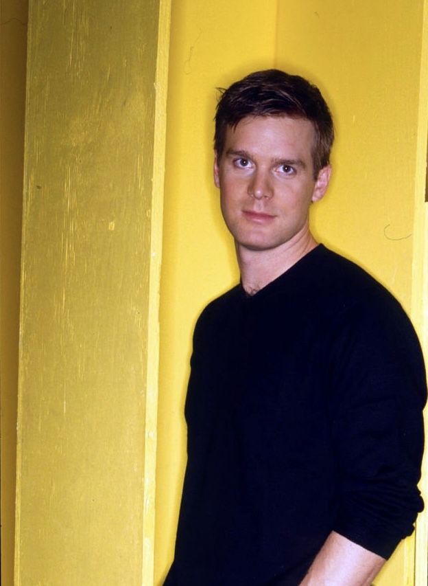 a man standing in front of a yellow wall with his hands in his pockets and looking at the camera