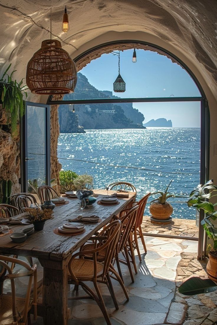 an outdoor dining area overlooking the ocean with chairs and table set for eight, in front of large arched window