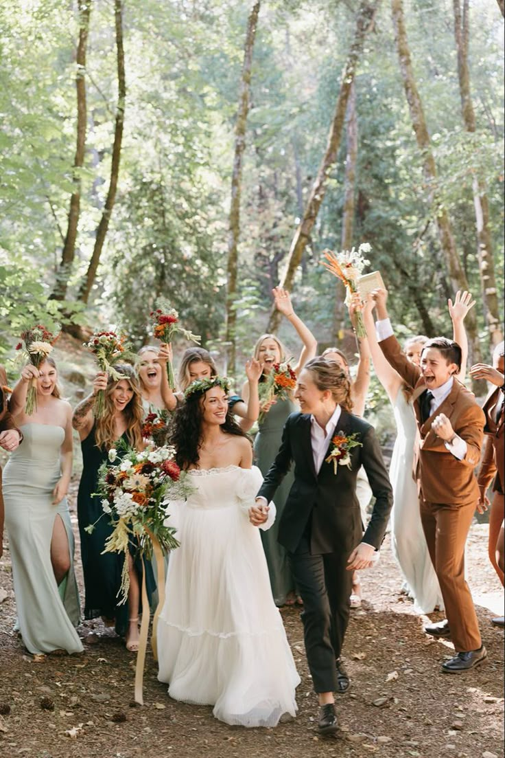 a bride and groom walking through the woods with their bridal party waving at them