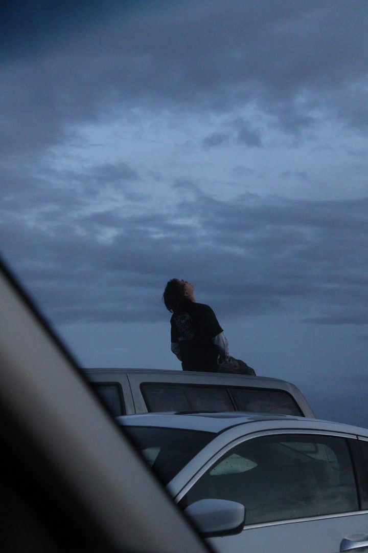 a man sitting on top of a car in front of some parked cars at dusk