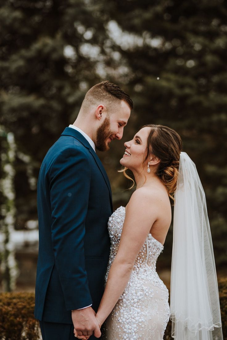 a bride and groom smile at each other while standing in front of some evergreen trees