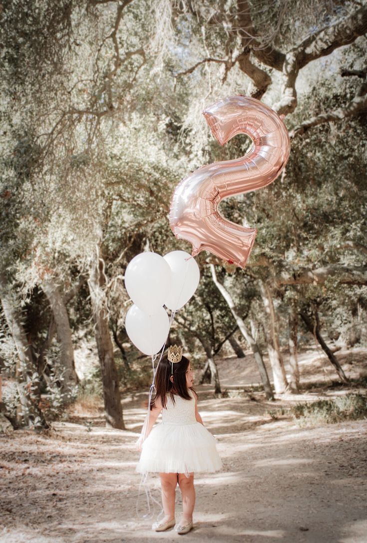 a girl in a white dress is holding balloons with the letter s on it and standing under some trees