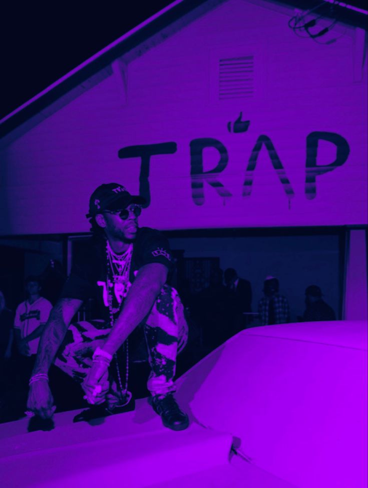 a man sitting on top of a car in front of a neon purple sign that says trap