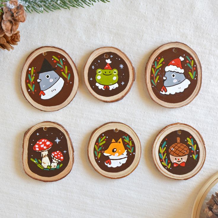 six decorated wooden magnets on a table with pine cones and fir tree branches in the background