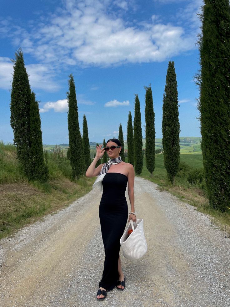 a woman walking down a dirt road holding a white bag in her hand and wearing sunglasses