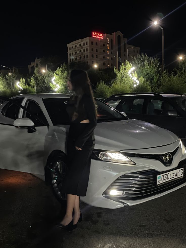 a woman standing next to a car in a parking lot at night with lights on