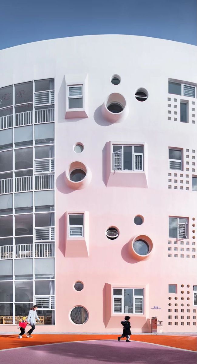 two people walking past a pink building with holes in the side and windows on it