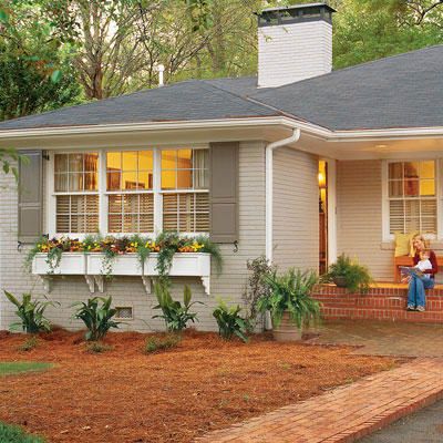 a small white house with trees and bushes around the front door, and a woman standing outside