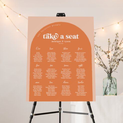 an orange and white wedding seating chart on a easel with lights in the background