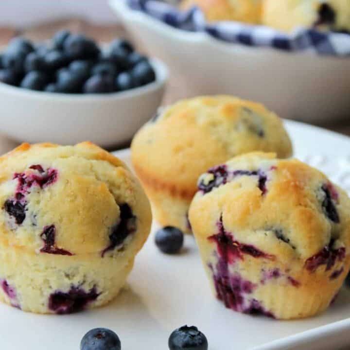 blueberry muffins on a white plate with bowls of blueberries in the background