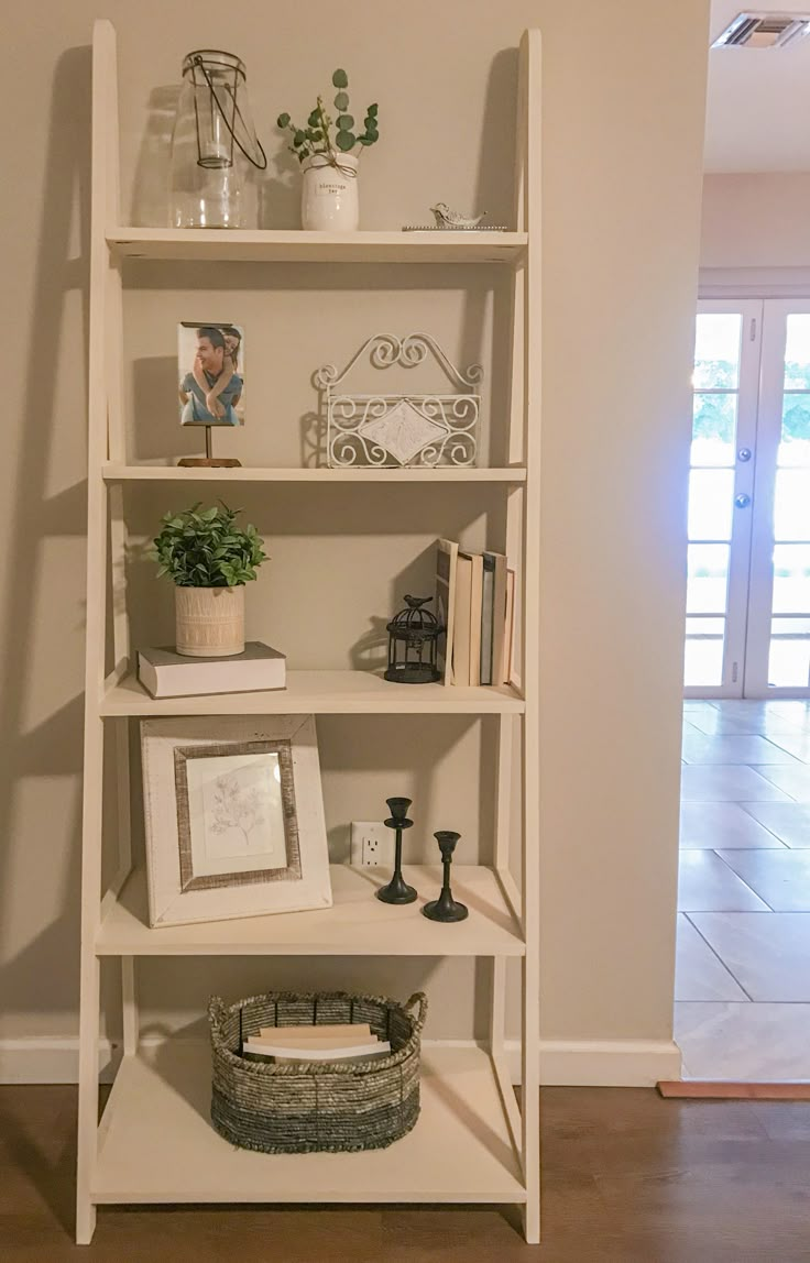 a white shelf with pictures and plants on it in the corner of a living room