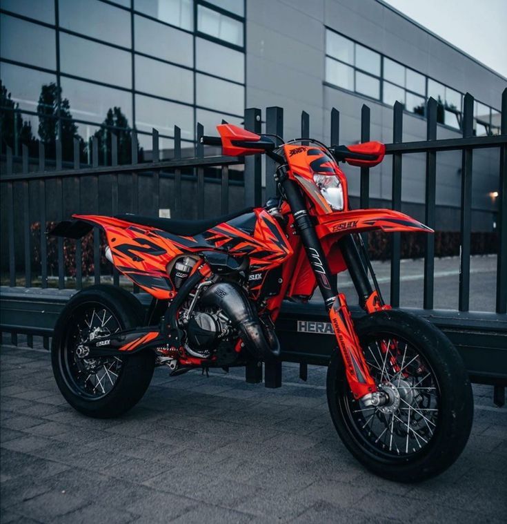 an orange and black dirt bike parked next to a metal fence in front of a building