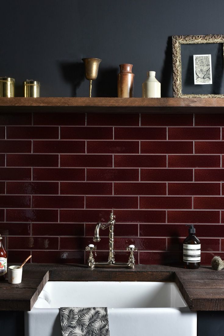 a kitchen sink and counter with red tiles on the wall behind it, in front of a wooden shelf