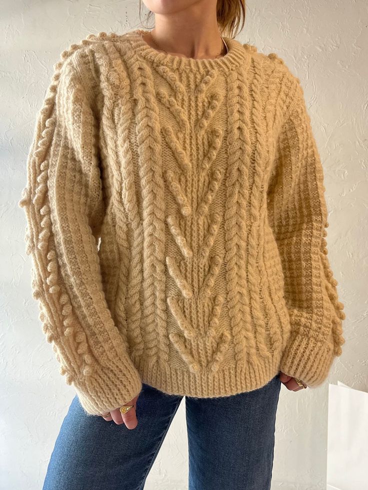 a woman wearing a beige sweater and jeans stands in front of a white wall with her hands on her hips