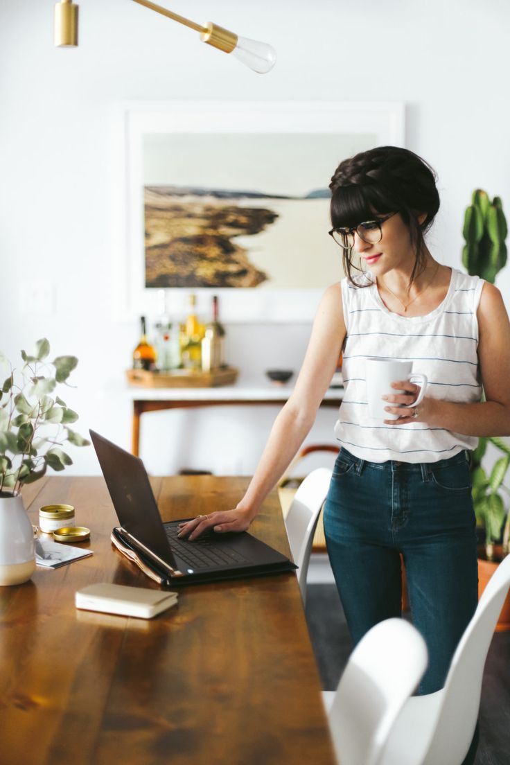 a woman sitting at a table with a laptop and coffee in her hand while looking at the screen