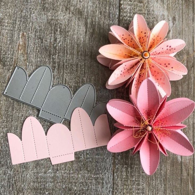 three paper flowers are placed next to each other on a wooden surface, one is pink and the other is gray