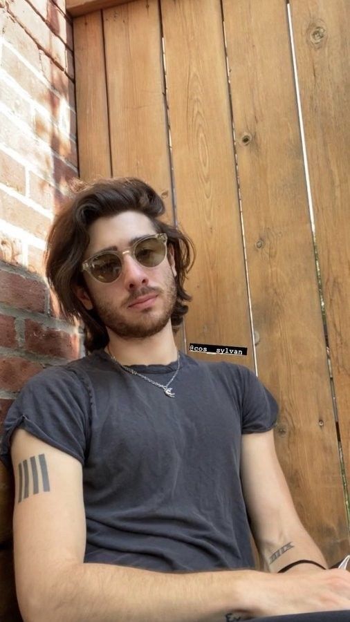 a man with long hair and sunglasses sitting on the ground next to a brick wall