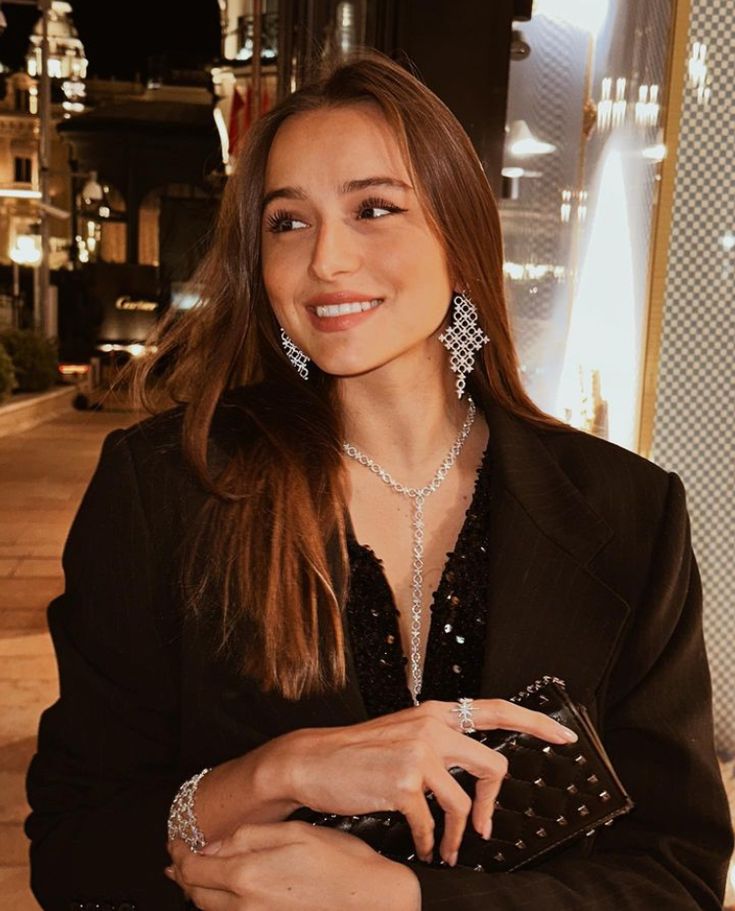 a woman in a black suit is holding a purse and smiling at the camera with her hand on her hip