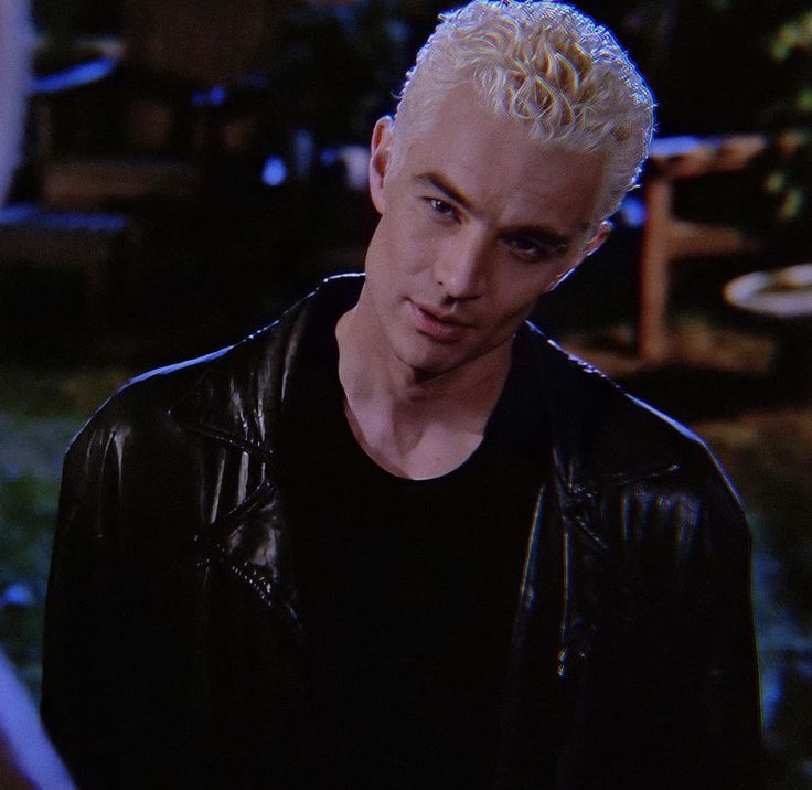 a man with blonde hair wearing a black jacket and looking at the camera while standing in front of a house