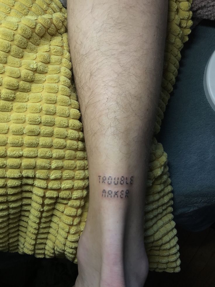 a man's foot with the words trouble maker tattooed on his left leg, sitting next to a yellow towel