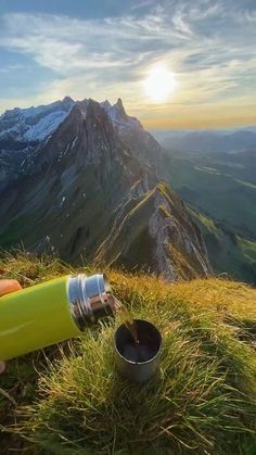 a person holding a water bottle on top of a grass covered hill with mountains in the background