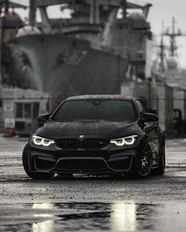 a black car is parked in front of a large ship and it's reflection on the wet ground