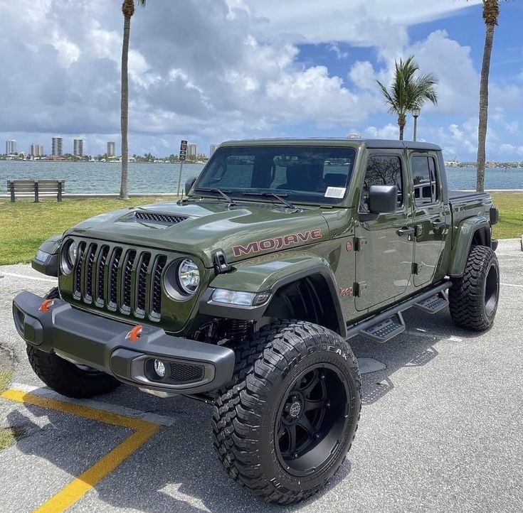 a green jeep parked in a parking lot next to the ocean and palm trees on a cloudy day