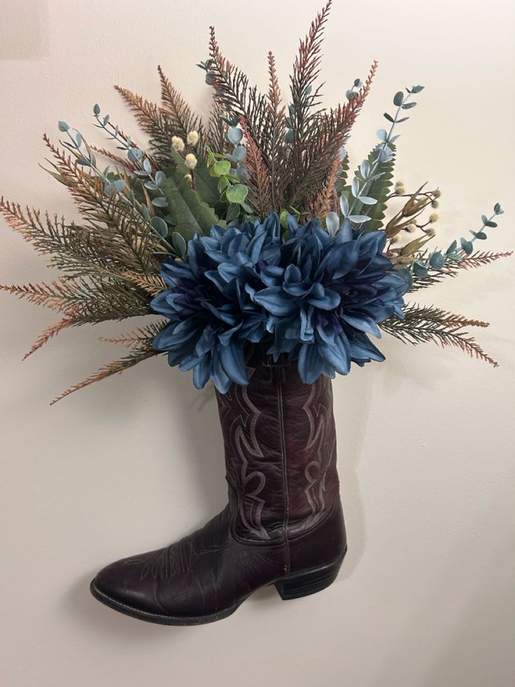 a cowboy boot with blue flowers and greenery in it hanging on the wall next to a white wall