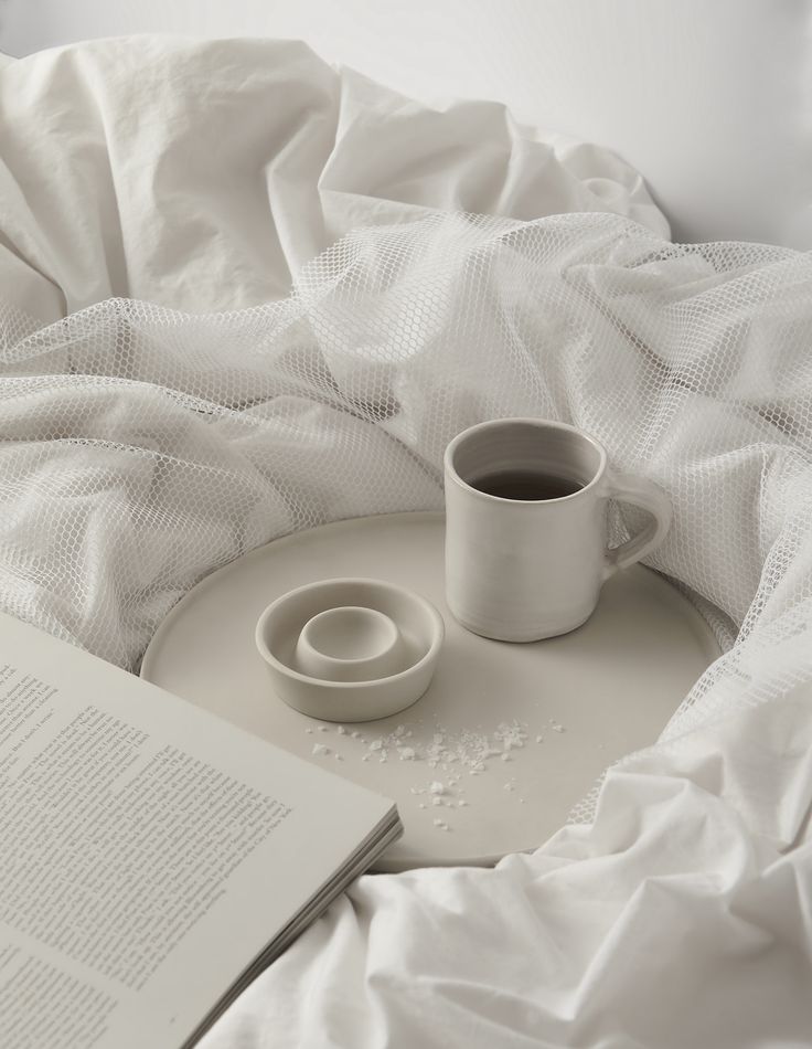 an open book and two mugs on a bed with white sheets in the background