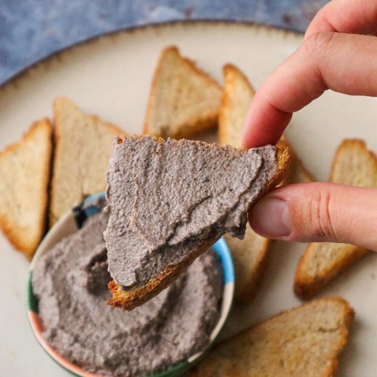 a hand holding a piece of bread over a bowl of hummus and crackers