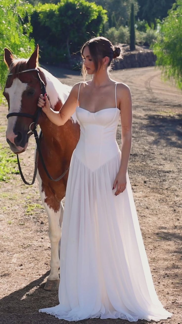 a woman in a white dress standing next to a brown and white horse on a dirt road