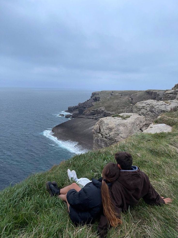 two people are sitting on the grass by the water looking out at the ocean and cliffs