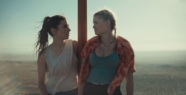 two young women standing next to each other on top of a hill looking at each other