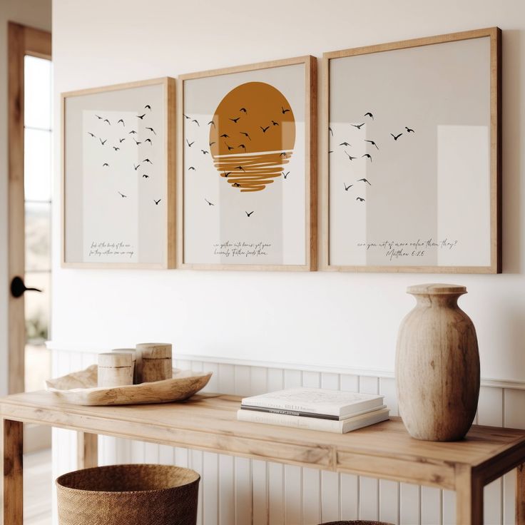 three framed pictures hang on the wall above a wooden table with a vase and book