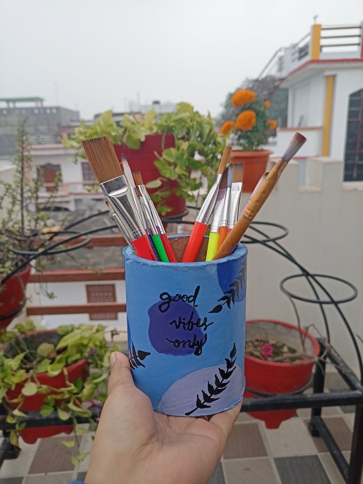 a hand holding a cup filled with lots of paint and pencils on top of a roof