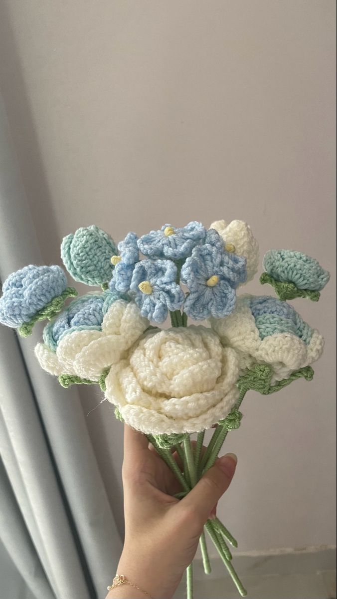 a hand holding a crocheted bouquet of blue and white flowers in front of a curtain