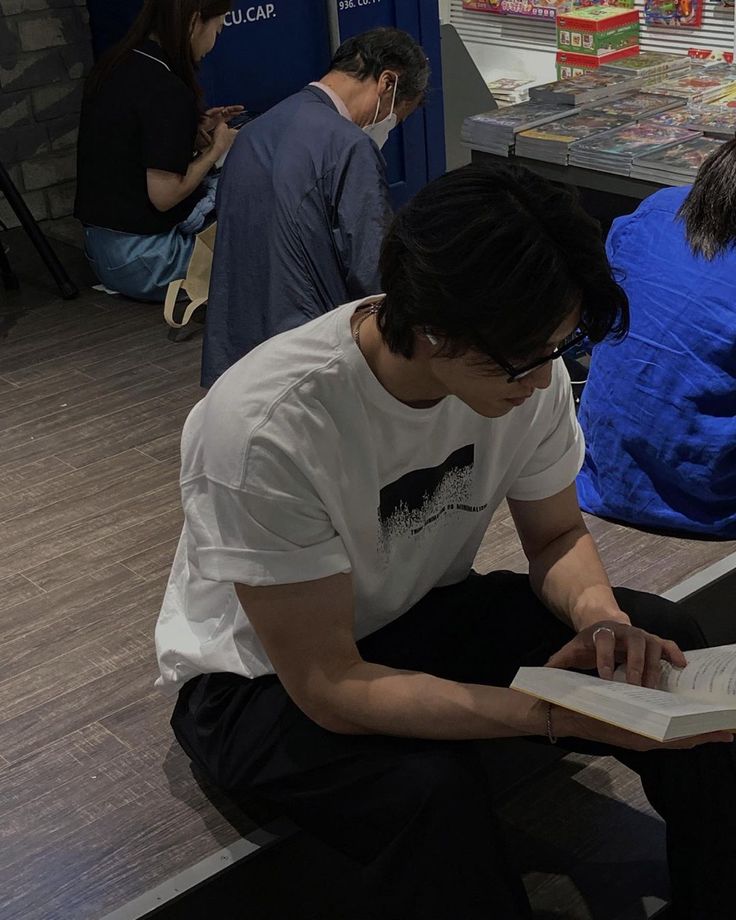 a young man sitting on the floor reading a book while others look at him in the background