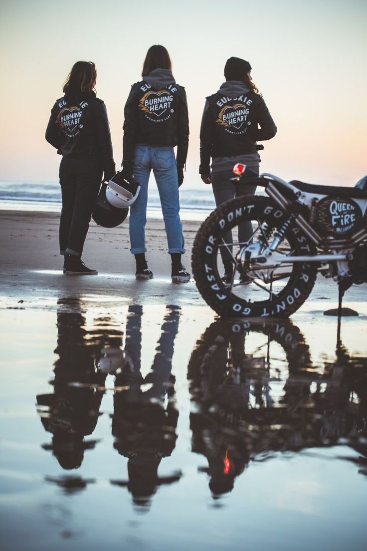 three people standing next to a motorcycle on the beach with their reflection in the wet sand