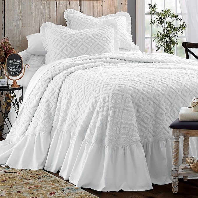 a bed with white ruffled bedspread and pillows on it in a bedroom