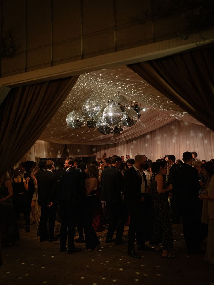 a large group of people standing around in a room with lights on the ceiling and drapes