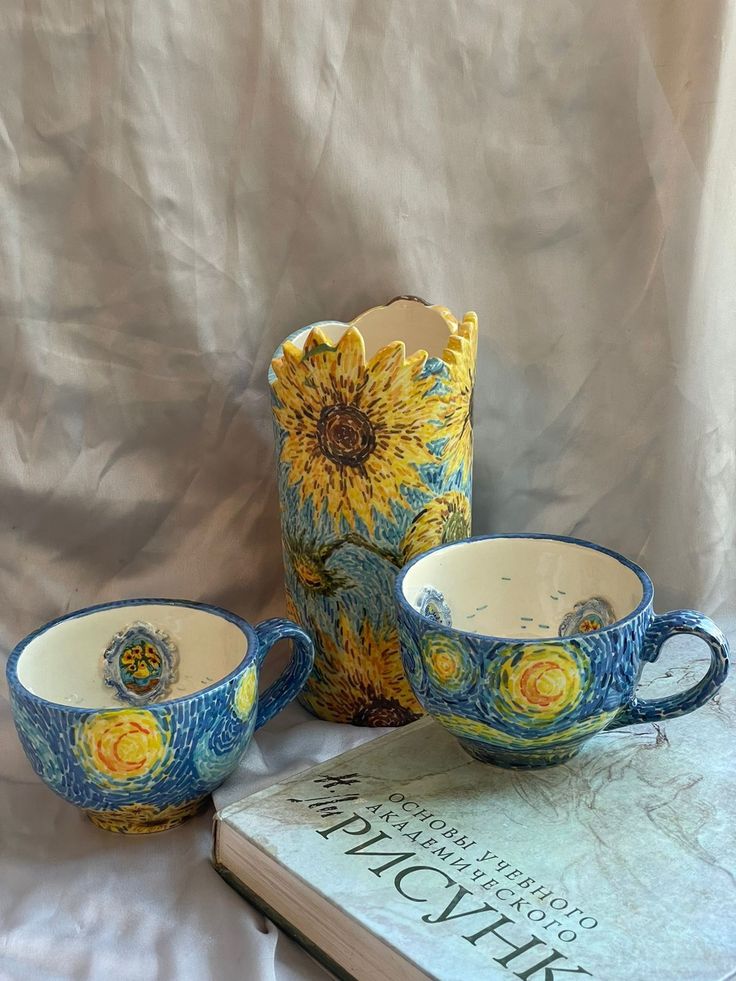 three cups and a vase sitting on top of a white cloth covered table next to a book