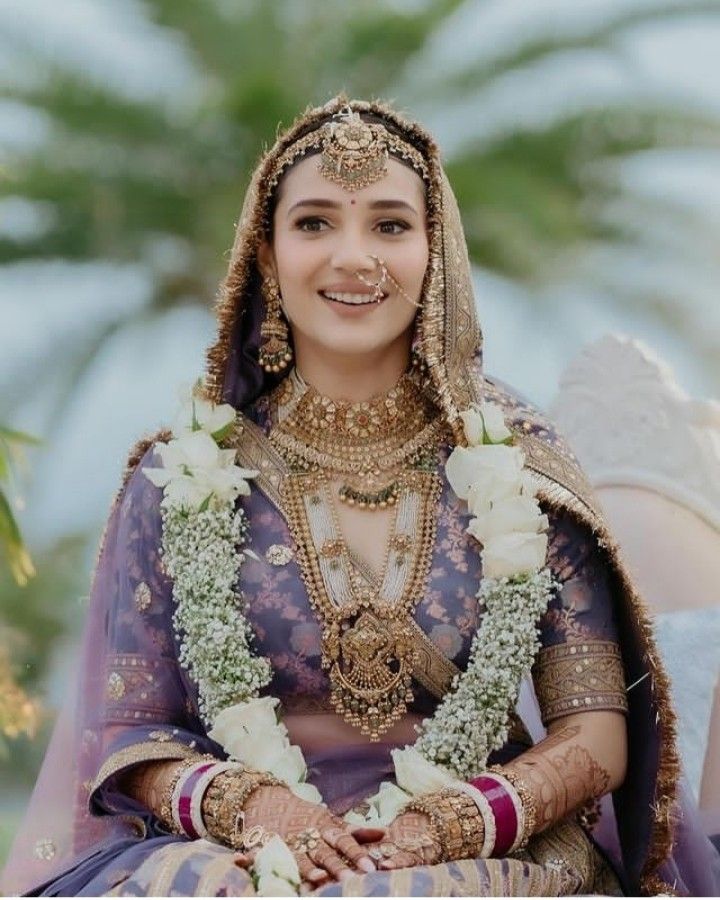 a woman in a purple and gold bridal outfit sitting on a white couch with flowers around her neck