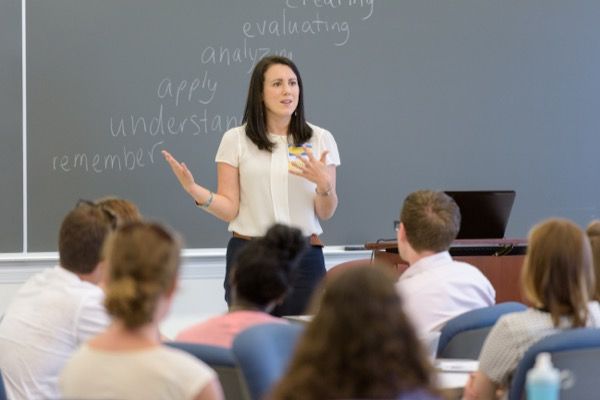 a woman standing in front of a class room full of students and giving a lecture